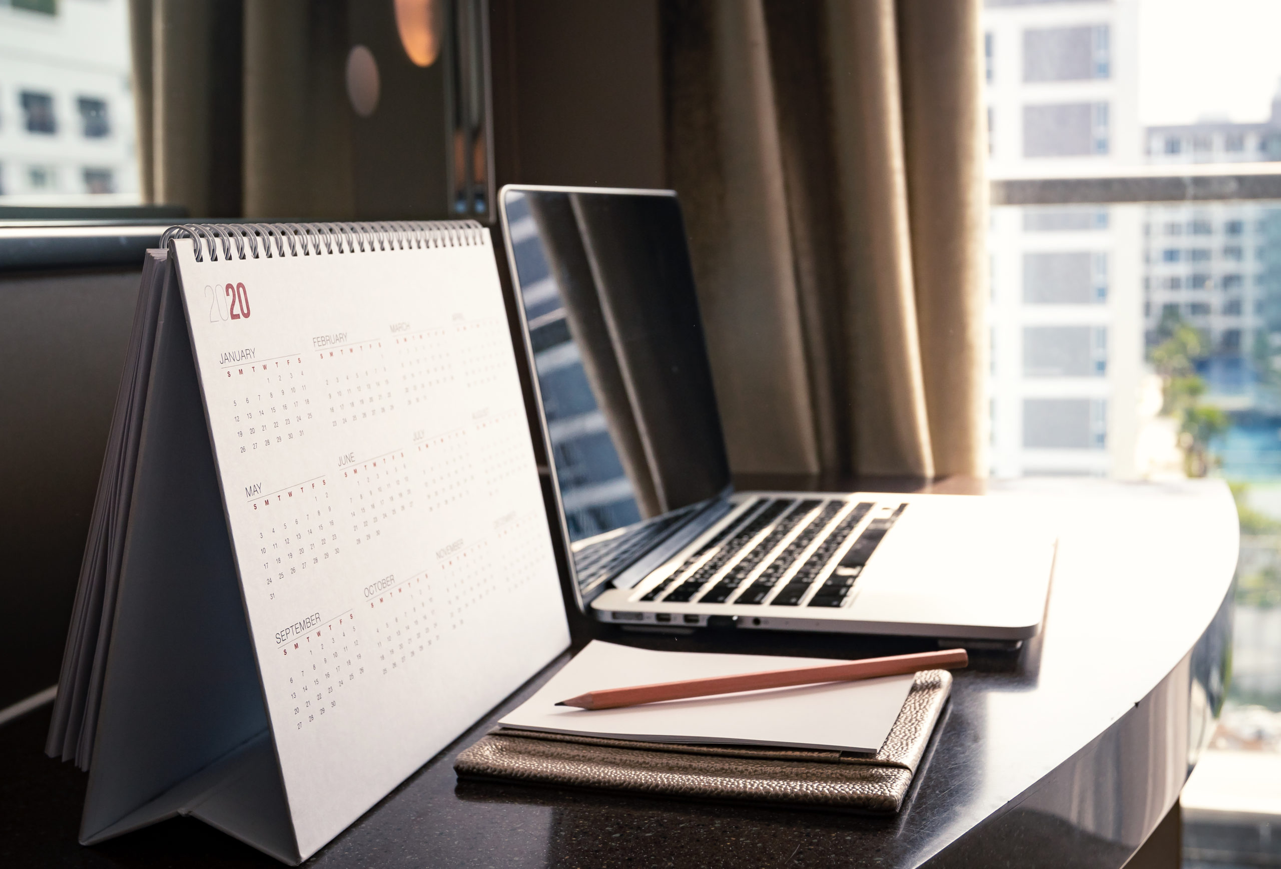 A calendar, computer, and notebook with pencil lay on top of a desk.