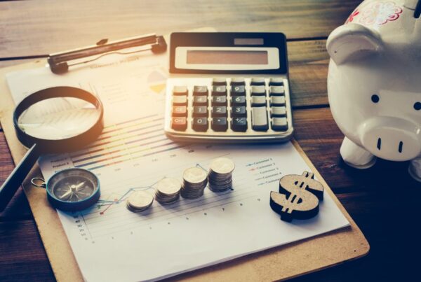 A calculator, stacks of coins, a compass, magnifying glass sit on a clipboard next to a piggy bank.